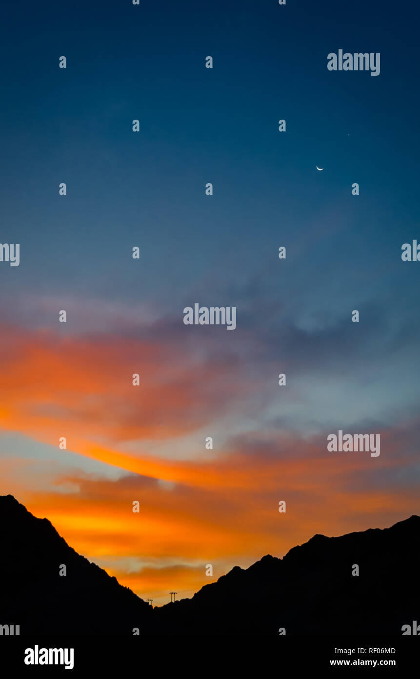 Shot of Orange clouds, blue sky, moon, star and silhouette of mountains in the morning. Stock Photo