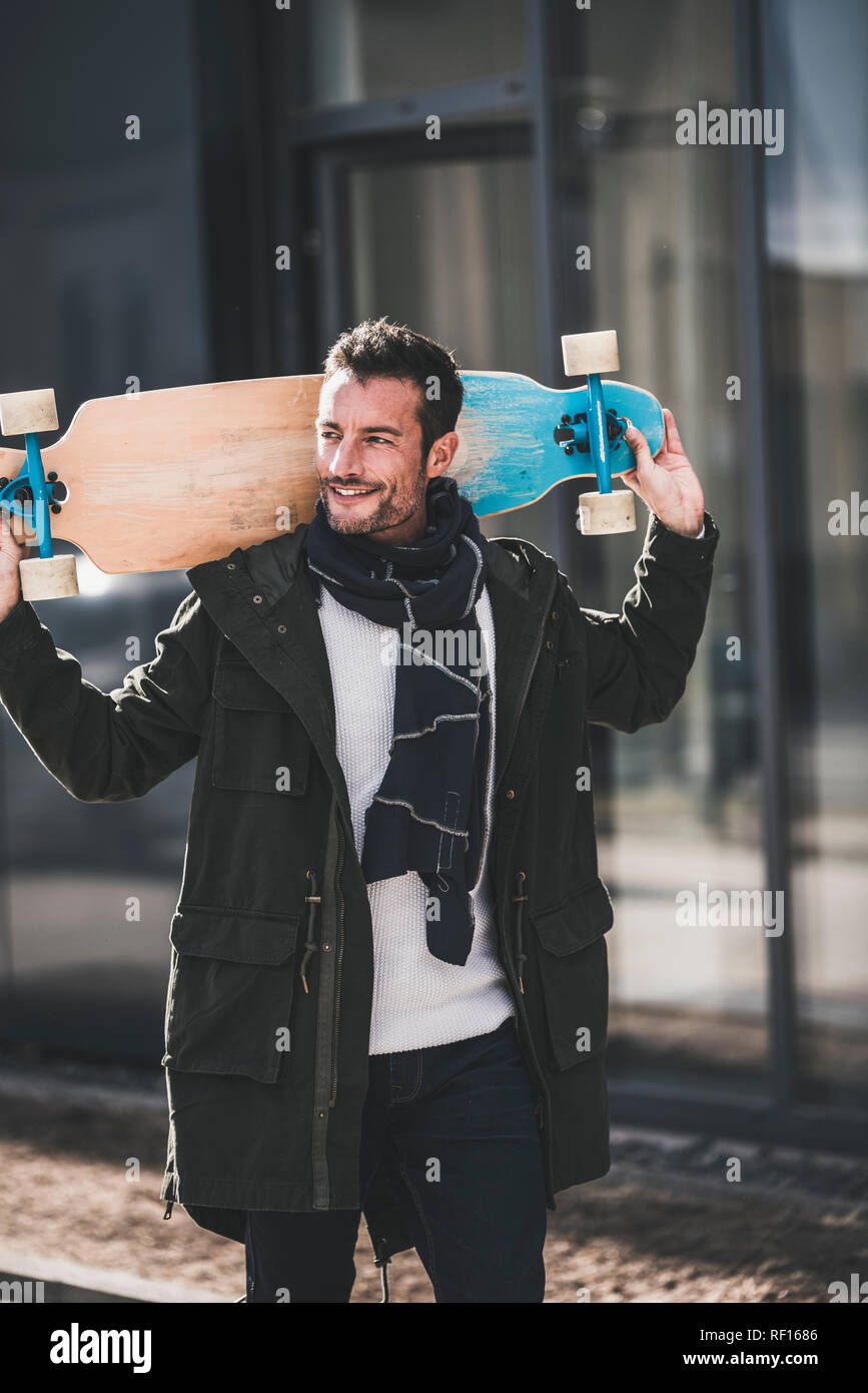 Mature man, wearing coat and scarf, walking in the city, carrying longboard Stock Photo