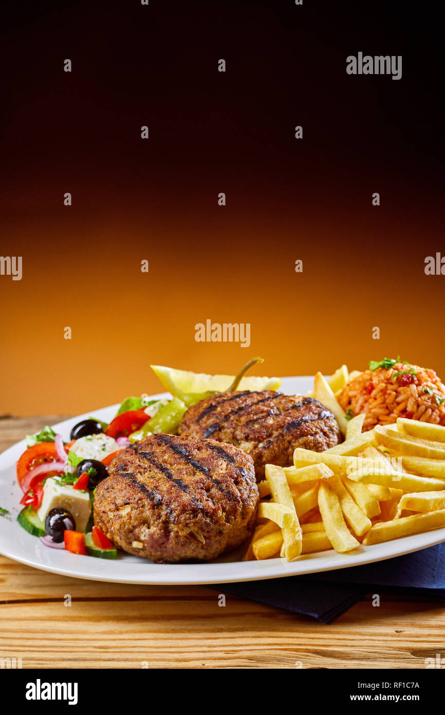 Grilled Greek bifteki, or spicy minced meat balls, fresh salad, domatorizo and French fries on a plate with copy space above Stock Photo