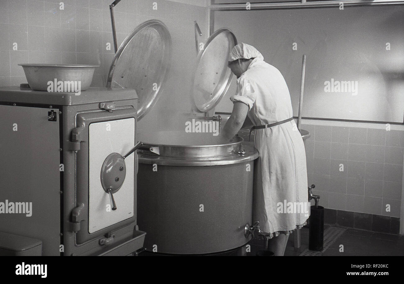 1950s, historical, steam rises from a mangle as a woman works in the laundry room of a county secondary school, England, UK. As can be seen, the equipment being used are eletric powered wringer or mangle type washing machines which squeezed the water out, hence the taps at the bottom. Large machines as these needed experienced handling. Stock Photo