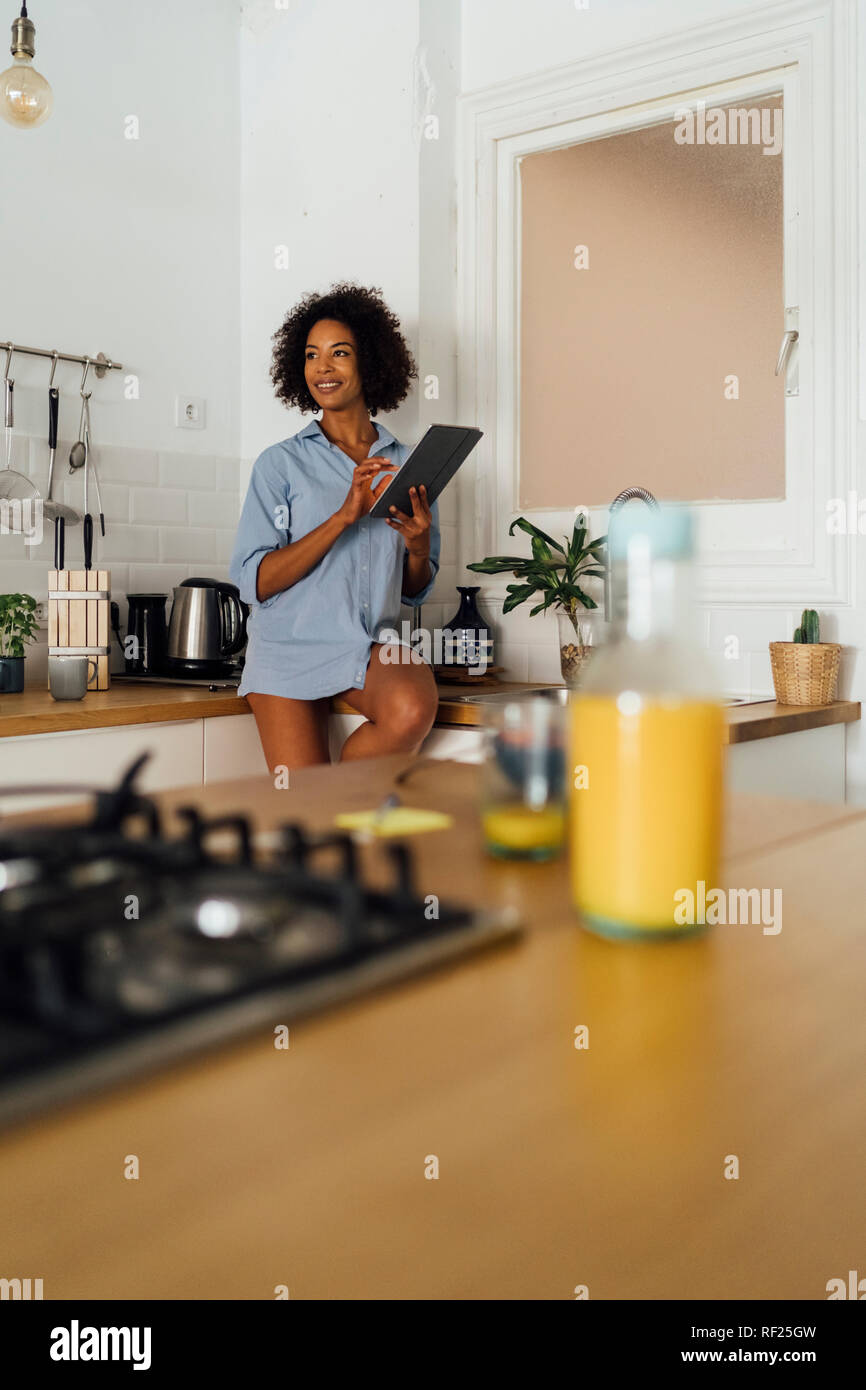 Woman sitting on worktop of her kitchen, using digital tablet in the morning Stock Photo