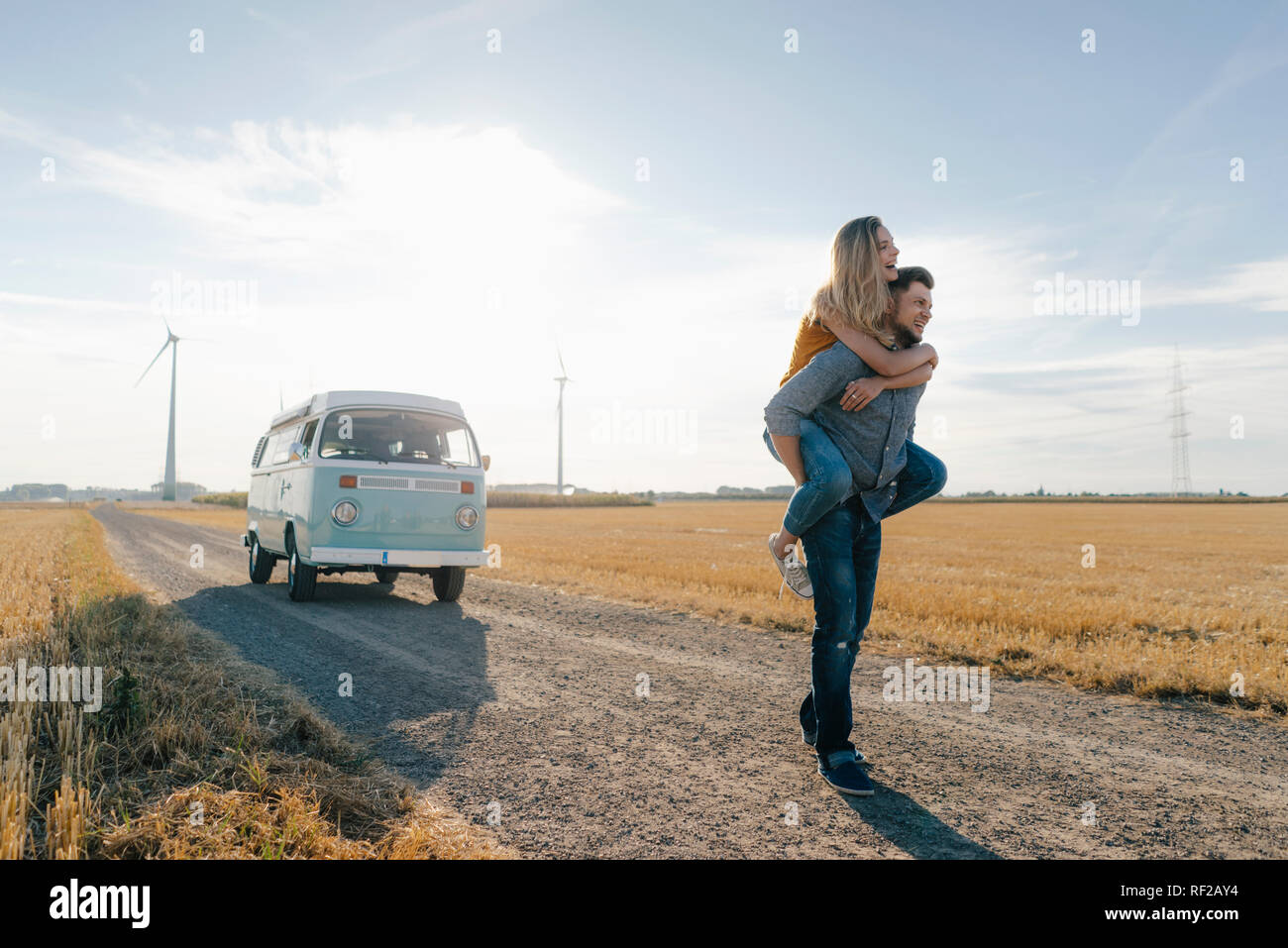 Young man carrying girlfriend piggyback on dirt track at camper van in rural landscape Stock Photo