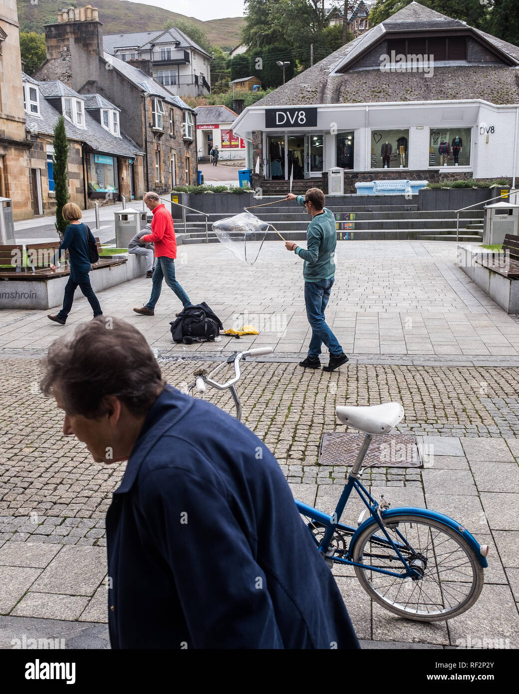 Old woman and couple walk past a parked blue bicycle and street performer in fort william high street highlands scotland uk Stock Photo