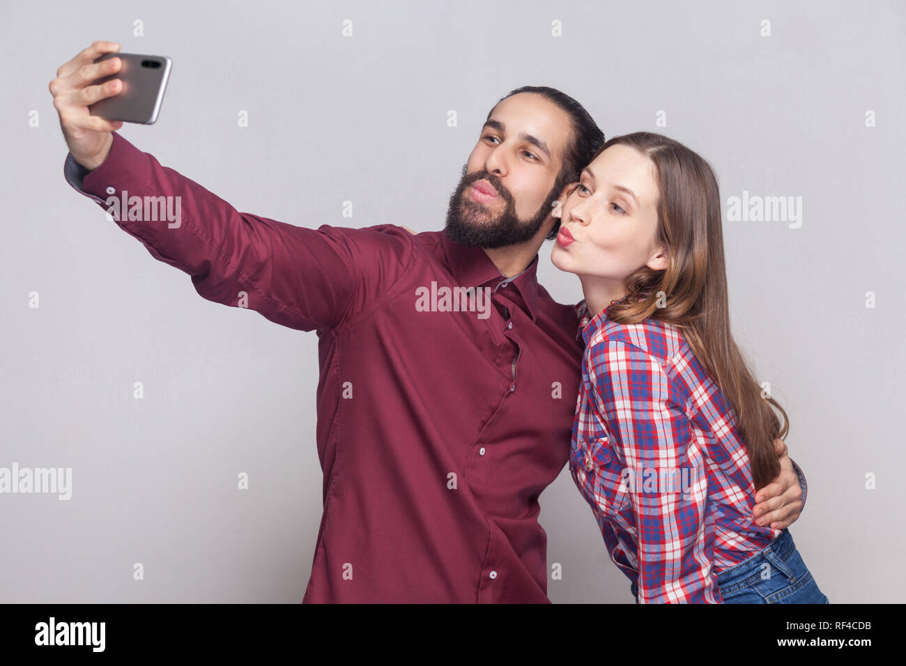 Portrait of happy couple standing, looking and kissing at camera of mobile smart phone to make selfie or video call. indoor studio shot, isolated on g Stock Photo