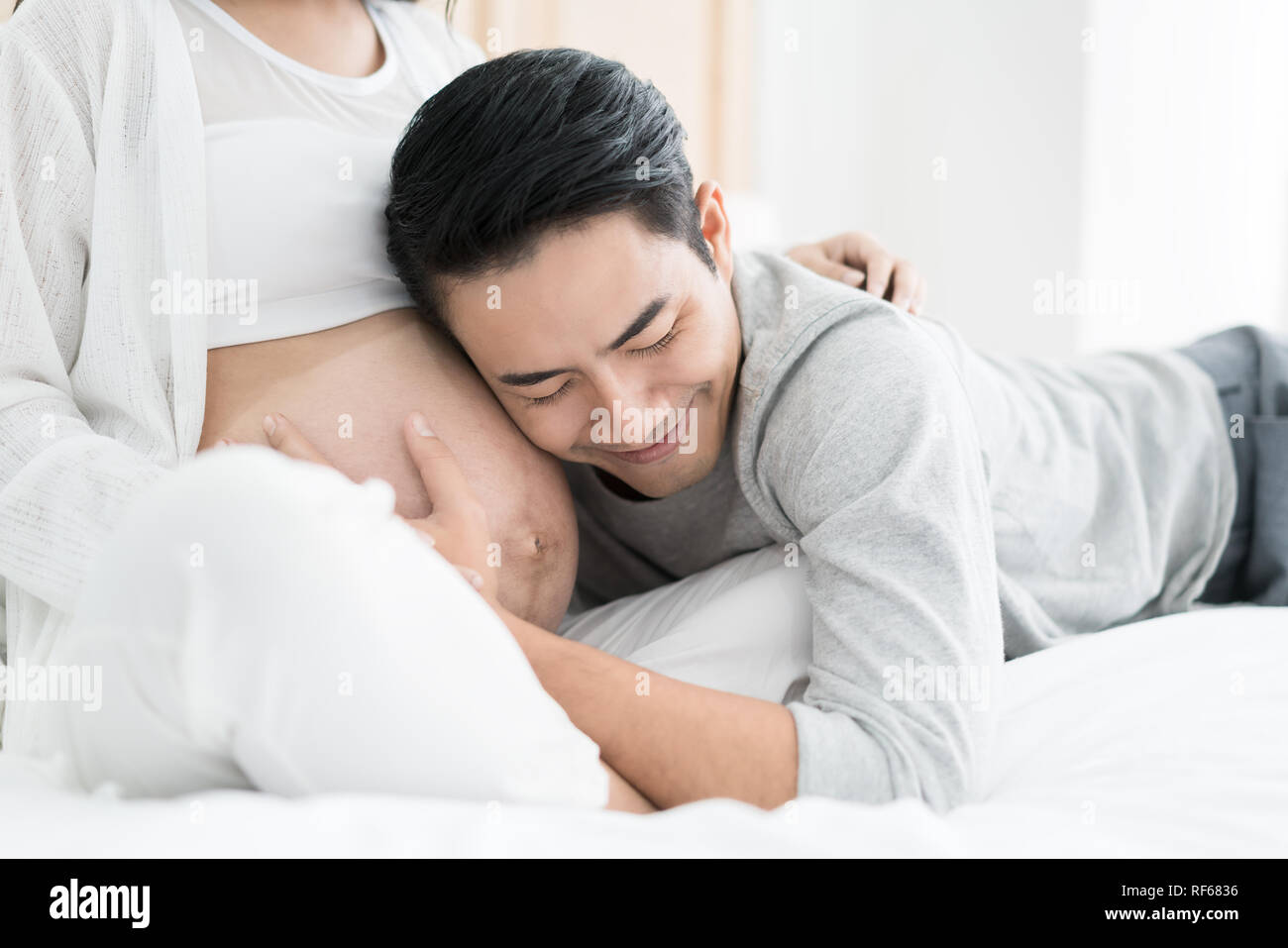 Asian Handsome man is listening to his Asian beautiful pregnant wife's tummy and smiling. Family love concept. Stock Photo