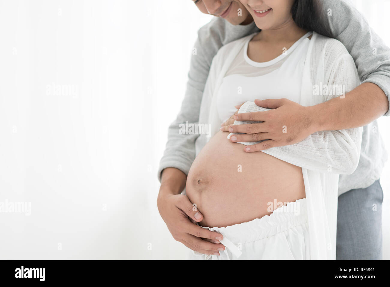 Handsome Asian man and his beautiful pregnant wife are hugging and smiling while standing near the window at home. Stock Photo