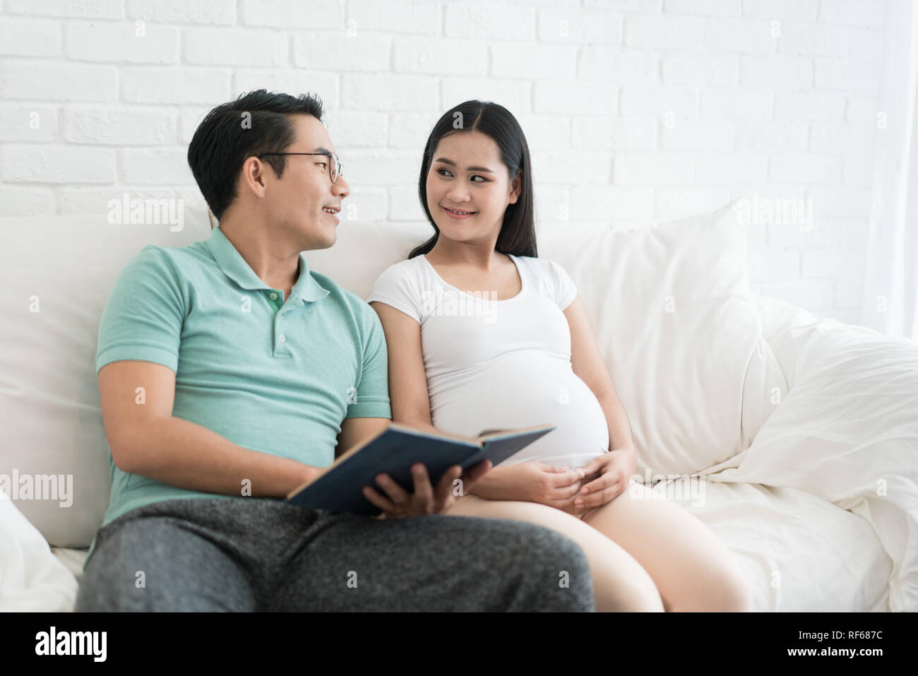 Close-up of Asian pregnant woman and Asian husband reading a book on the sofa at home. Happy family waiting newborn concept. Stock Photo