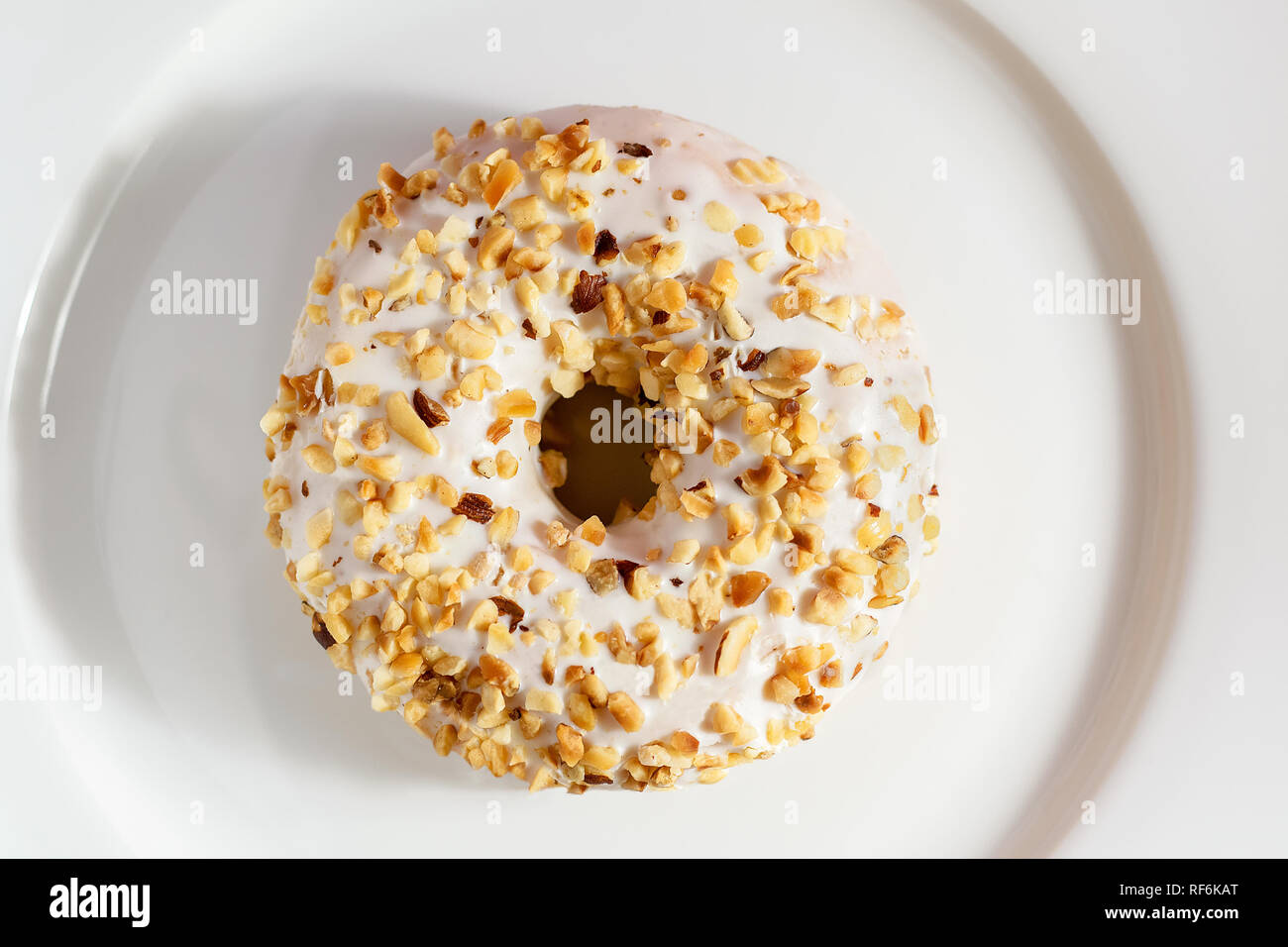 Donut with white chocolate and sprinkled with nuts on top, laid on white glossy plate. Stock Photo