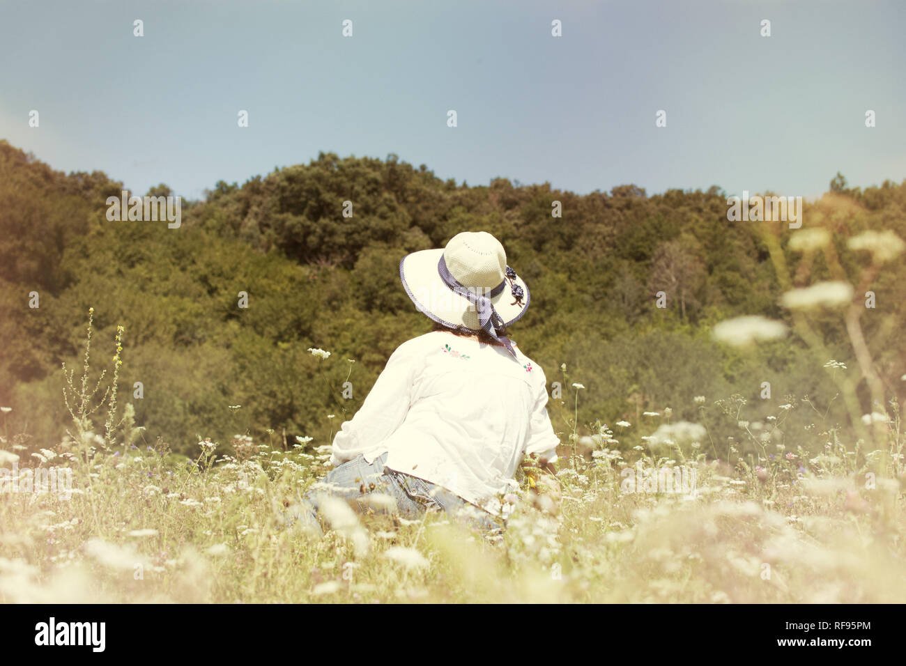 PORTRAIT OF BACWARDS SENIOR WOMAN SITTING ENJOING SPRING LANDSCAPE, WEARING A HAT. Stock Photo
