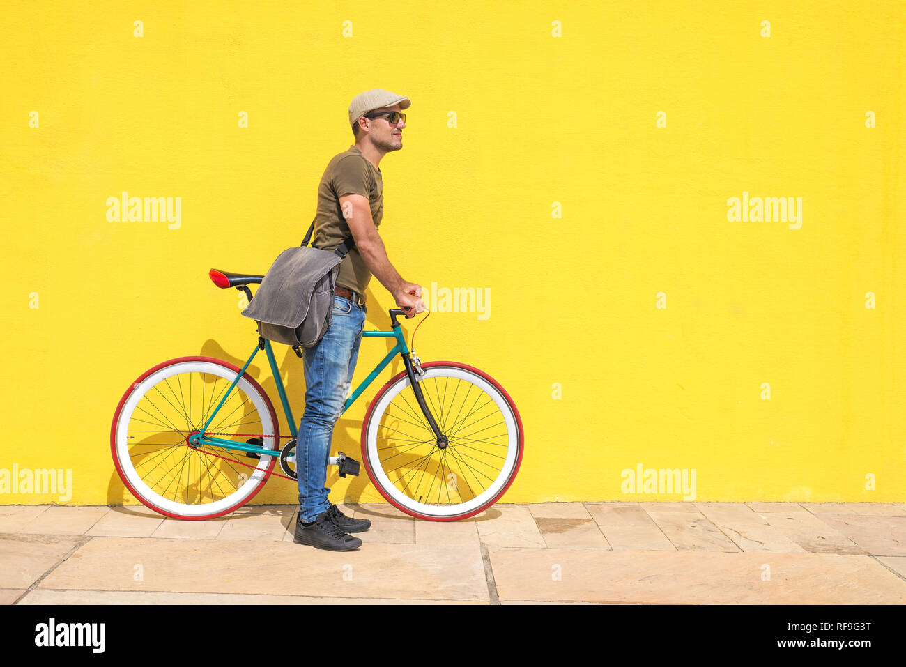 Side view of a young trendy man with a fixed bike wearing casual clothes while looking away against a yellow wall outdoors in a sunny day Stock Photo