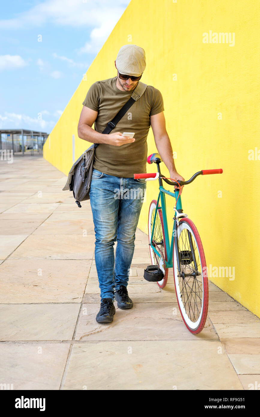 Front view of a young trendy man with a fixed bike wearing casual clothes while using a mobile phone against a yellow wall outdoors in a sunny day Stock Photo