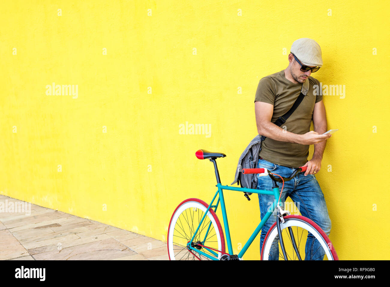 Side view of a young man with a vintage bike and wearing casual clothes and sunglasses standing against a yellow wall while using a mobile phone in a  Stock Photo