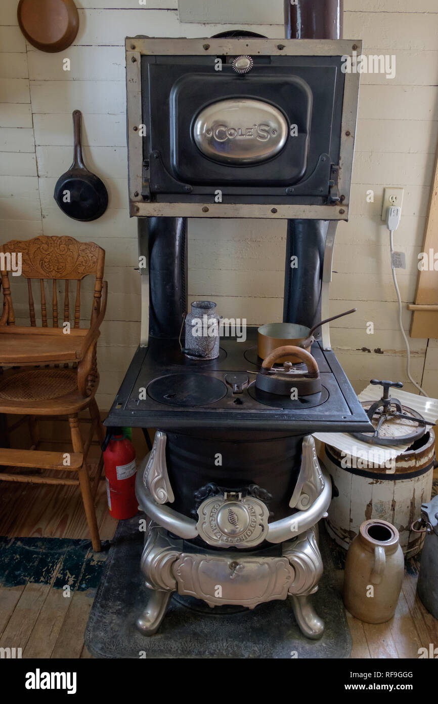 Vintage kitchen interior with antique Coles coal stove with oven, vintage cooking utensils, old wooden high chair, cast iron fry pan, earthen jug. Stock Photo