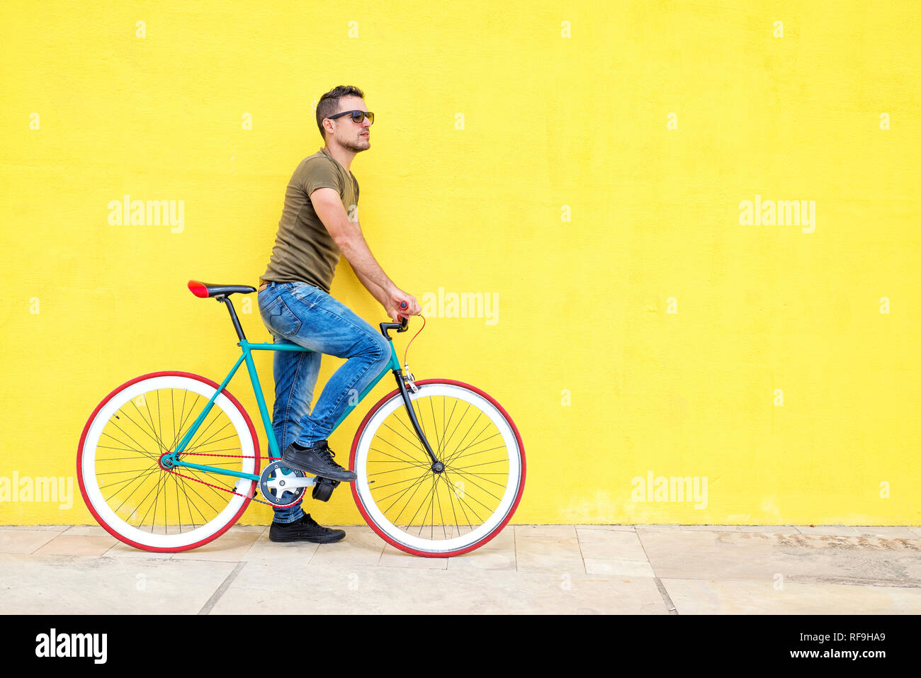 Side view of a young trendy man with a fixed bike wearing casual clothes while looking away against a yellow wall outdoors in a sunny day Stock Photo