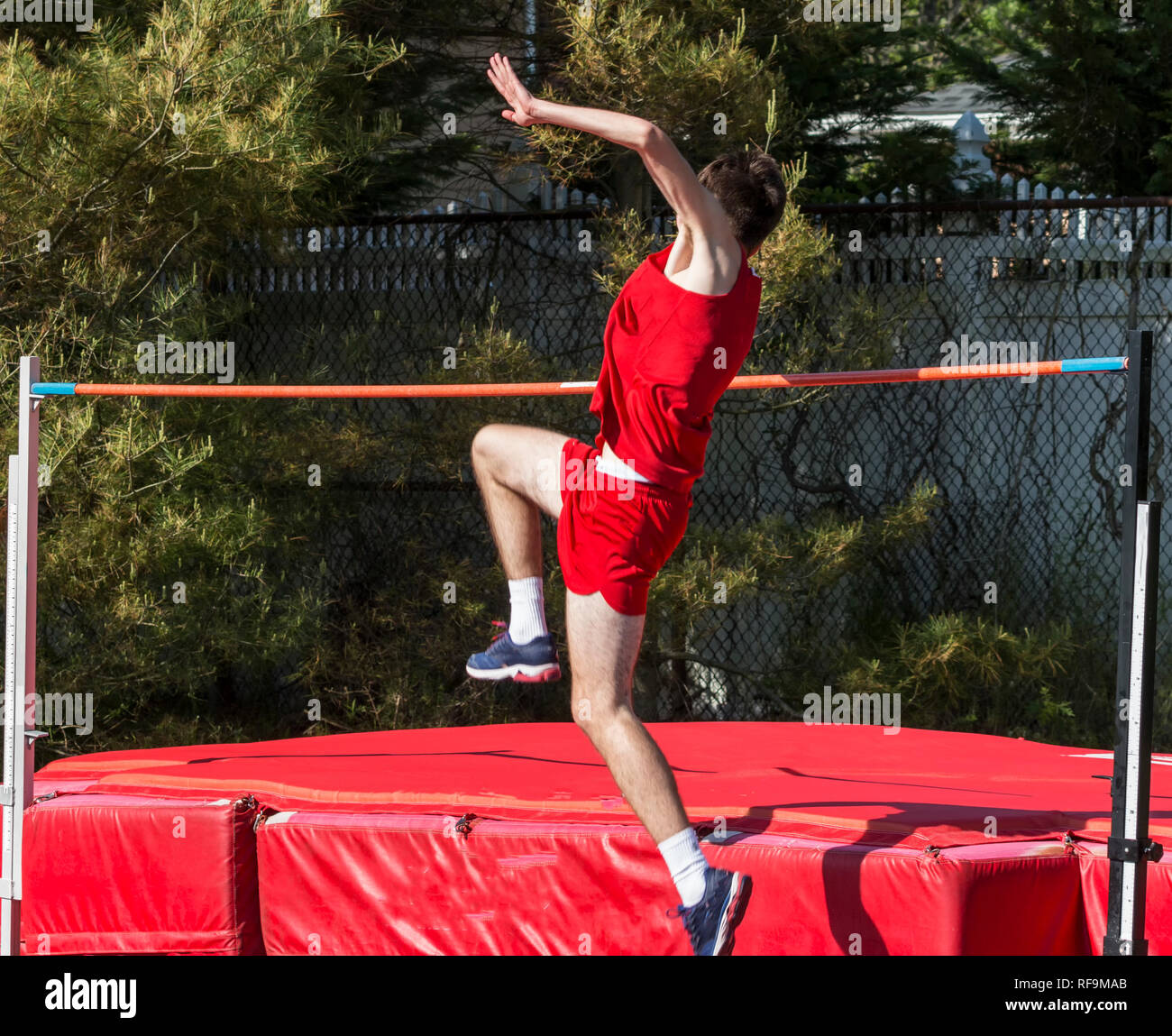 A high school track and field high jumper is taking off from the ground as he attempts to jump over the orange bar onto the red matts during a high ju Stock Photo