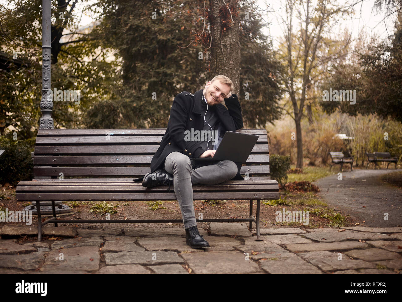 One Young Relaxed Man Sitting On Bench In Public Park Using