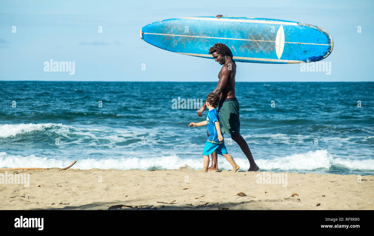 An athletic man carrying a blue surfboard and a young boy wearing blue clothes are walking on a sandy beach on the background of deep blue ocean. Stock Photo