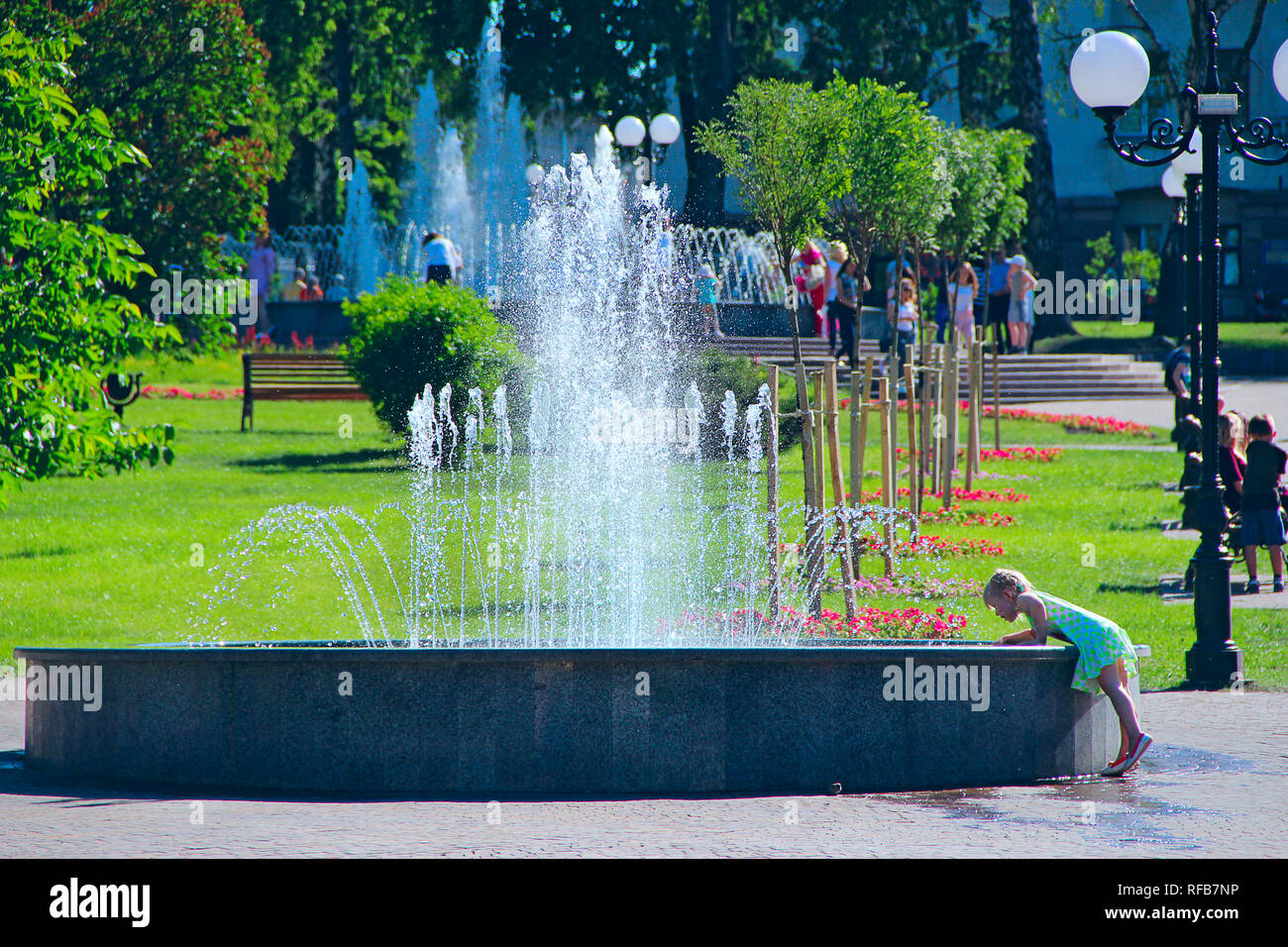 Little girl trying to touch water in city fountain. People have a rest in city park with fountains. Sunny summer day. Child playing with water of city Stock Photo