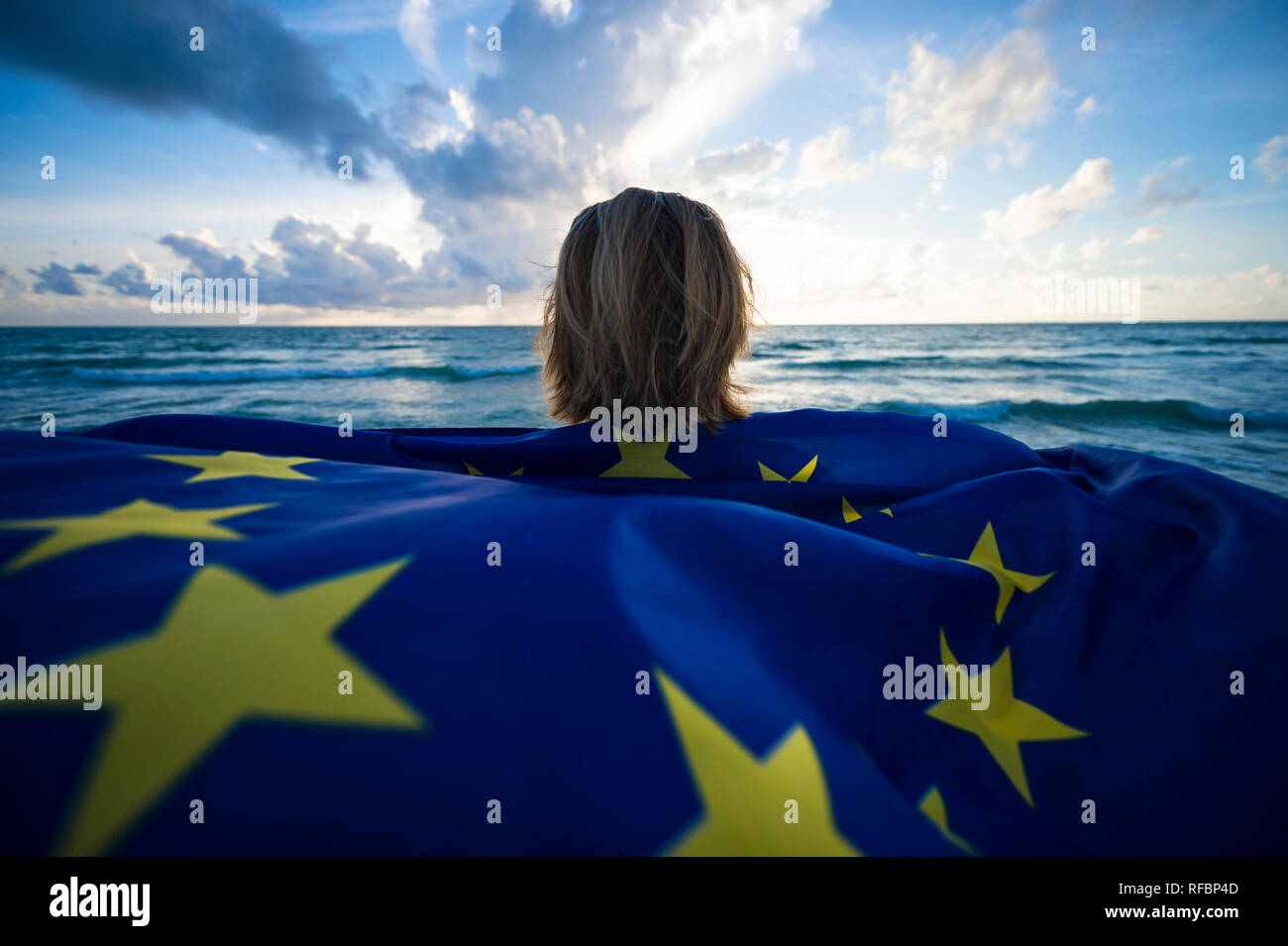 Man holding a fluttering iconic EU European Union flag with circle of stars on beach with stormy turbulent seas in the Channel at sunrise Stock Photo