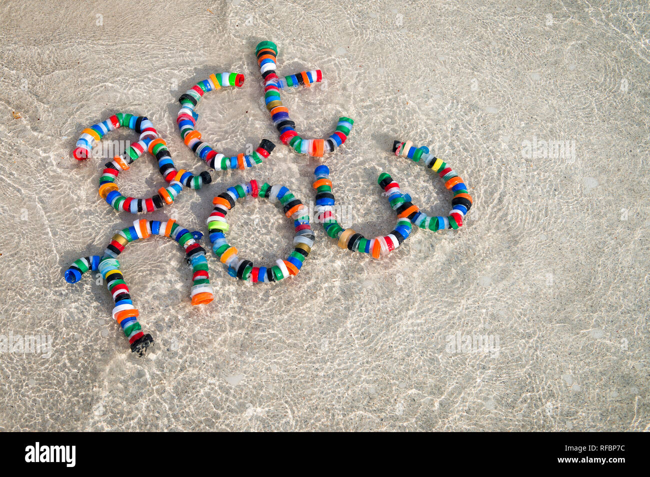 Colorful plastic bottle caps spell out 'act now' on a sandy beach with lapping waves, a reminder for people to take action, reduce, reuse and recycle, Stock Photo