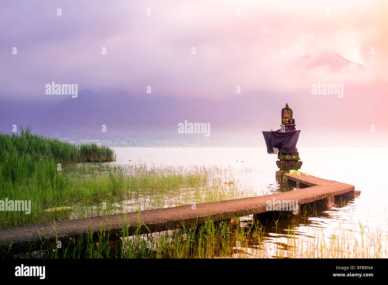 Small Hindu Temple on The Lake Side With Connecting Bridge During Sunrise in Bali on Cloudy and Misty Day with Mountain as Background. Stock Photo