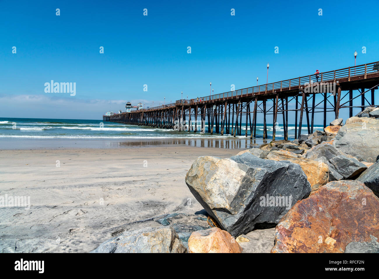 Pier running into sea, large boulders on right with beach and ocean beyond under bright blue sky. Stock Photo