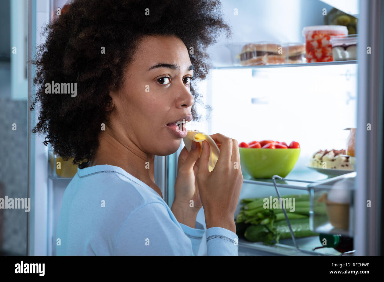 Young African Woman Eating Slice Of Cheese Near An Open Refrigerator Stock Photo