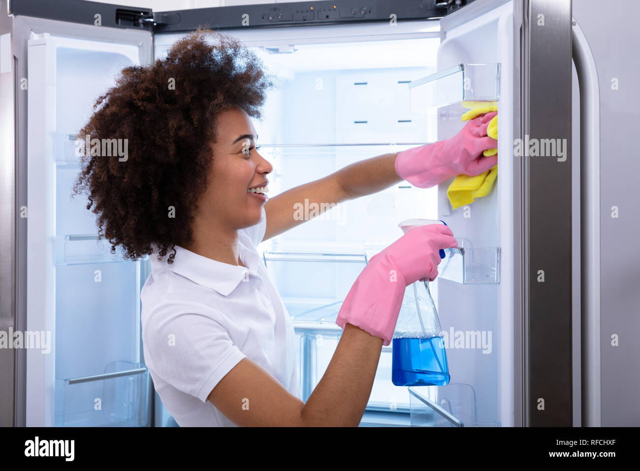 Young Happy Cleaning Lady Cleaning The Empty Refrigerator Door With Spray Bottle And Napkin Stock Photo