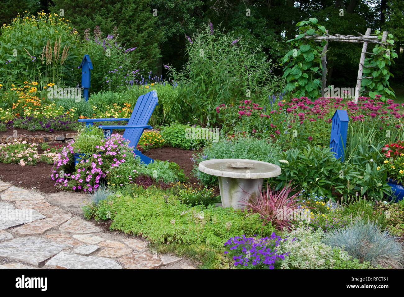 63821-21812 Flower garden with stone path, blue Adirondack chair, bird bath, butterfly house, bird house, and trellis, Purple Verbena, sedums, blue fe Stock Photo