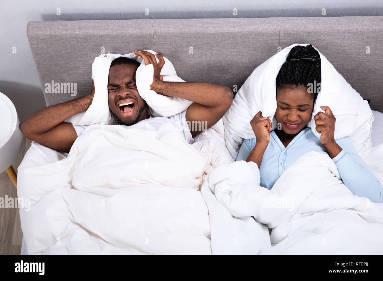 Young African Couple Disturbed By Noise Covering Their Ears With Pillow On Bed Stock Photo