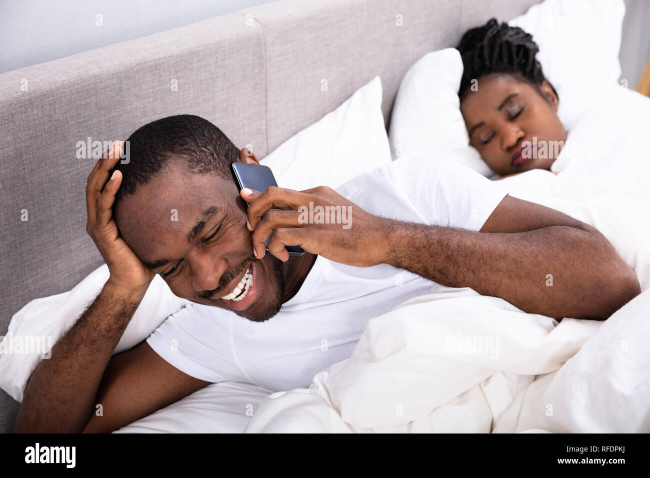 Smiling Young African Man Talking On Smartphone Besides His Wife Sleeping On Bed In Bedroom Stock Photo