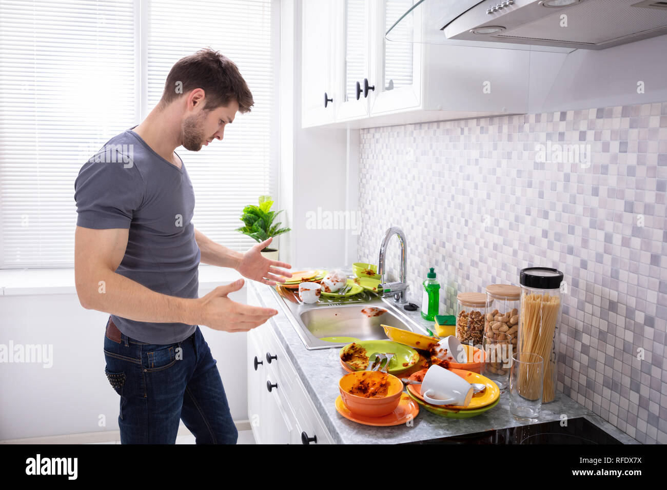 Sad Young Man Looking At Dirty Utensils Near Sink In Kitchen Stock Photo