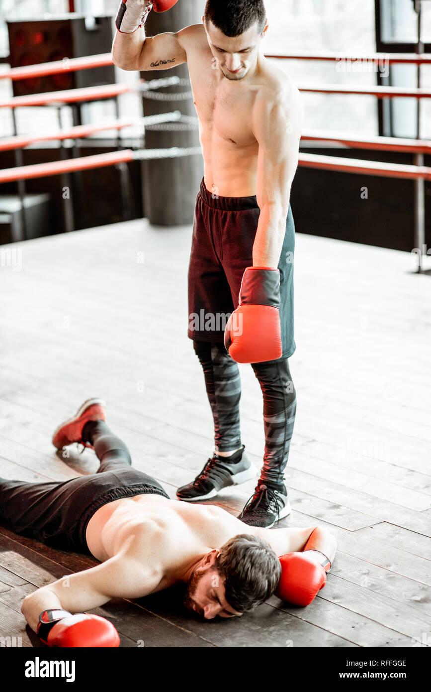 Beaten boxer lying knocked out on the boxing ring with strong man winner above Stock Photo