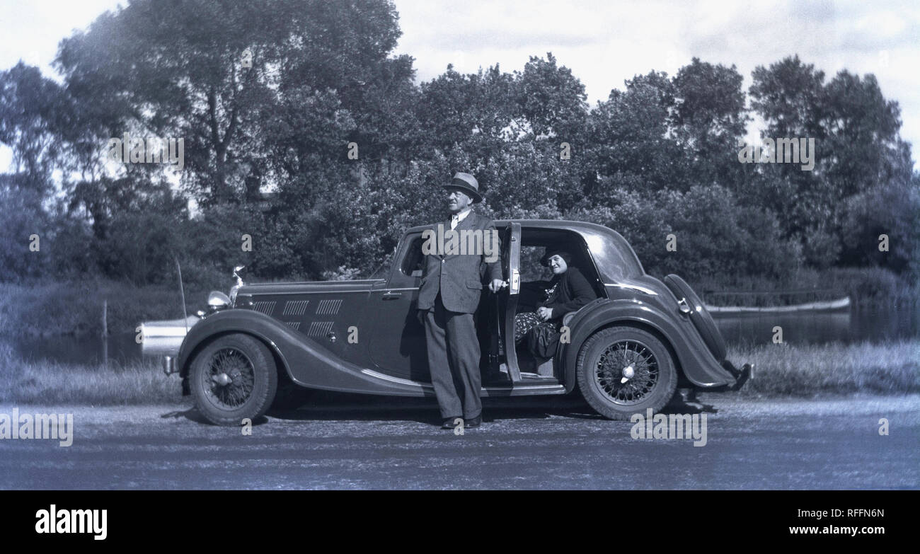 1930s, historical, by a river, a husband hold open the rear door of his sportscar as his wife sits in the back, England, UK. Stock Photo