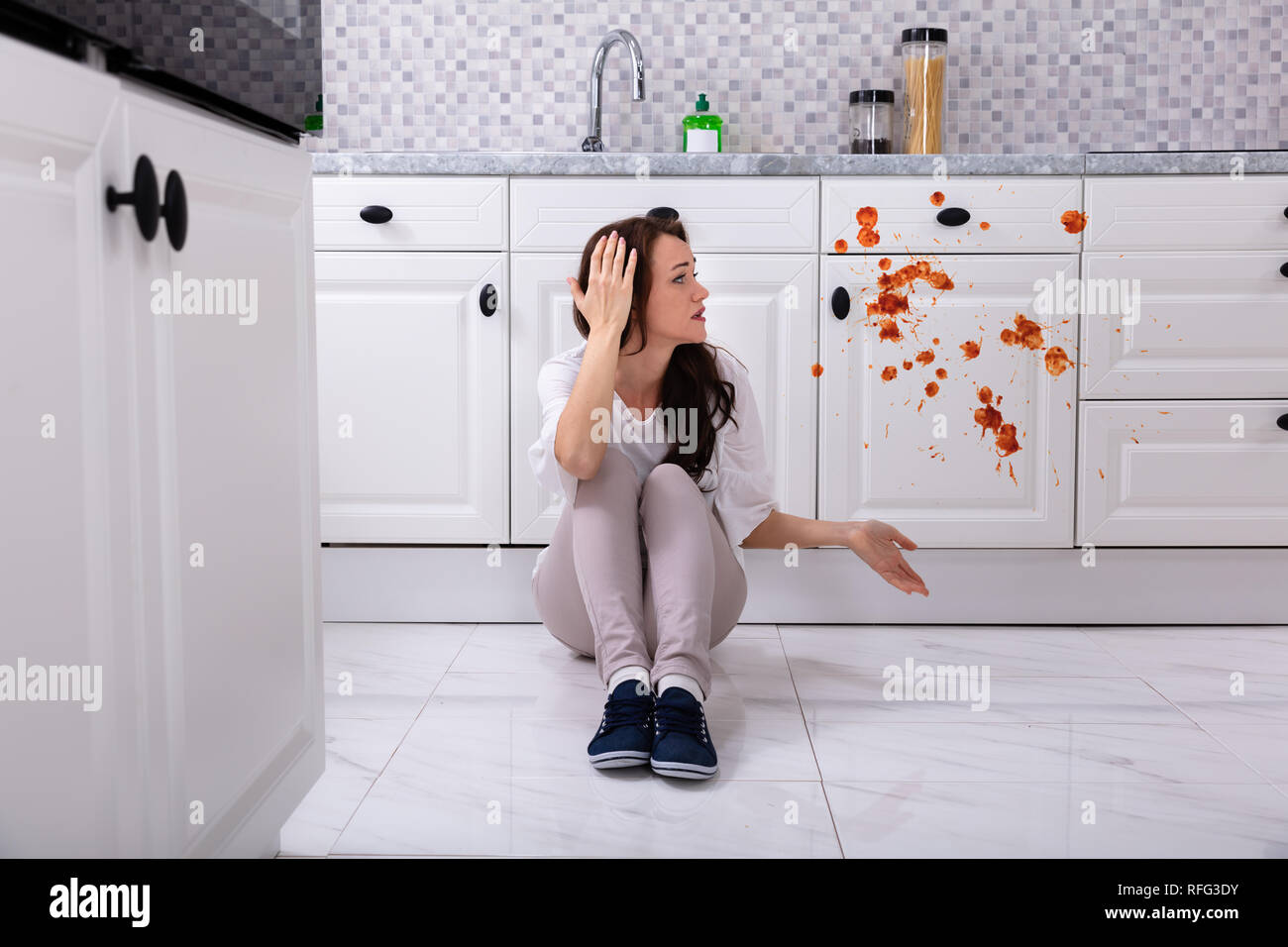 Sad Woman Sitting On Floor With Spilled Food In Kitchen Stock Photo
