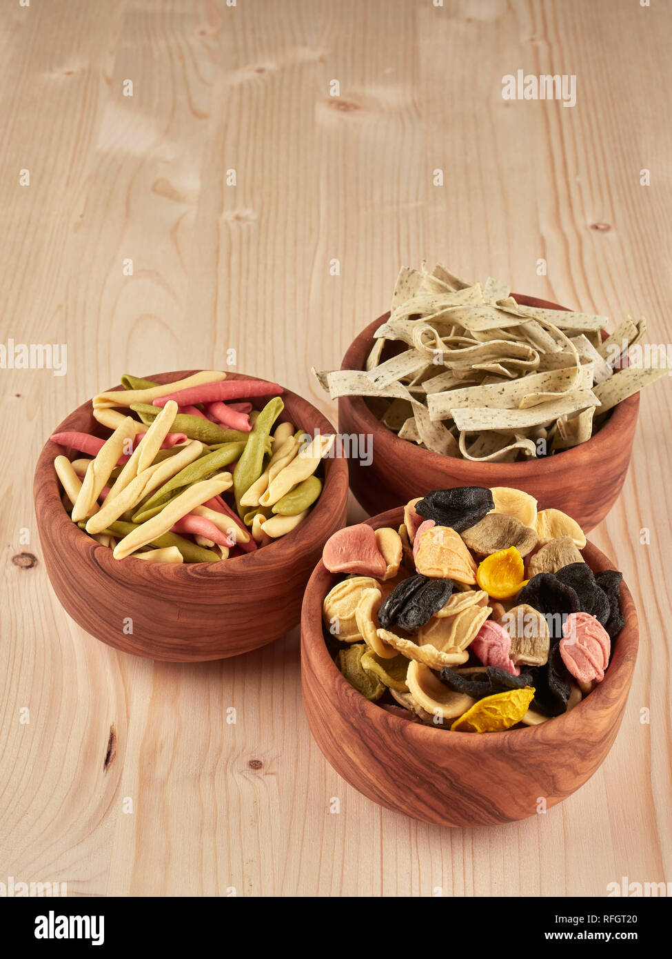 Three olive wood bowls with uncooked, colorful orechiette, fusilli and tagliatelle pasta Stock Photo