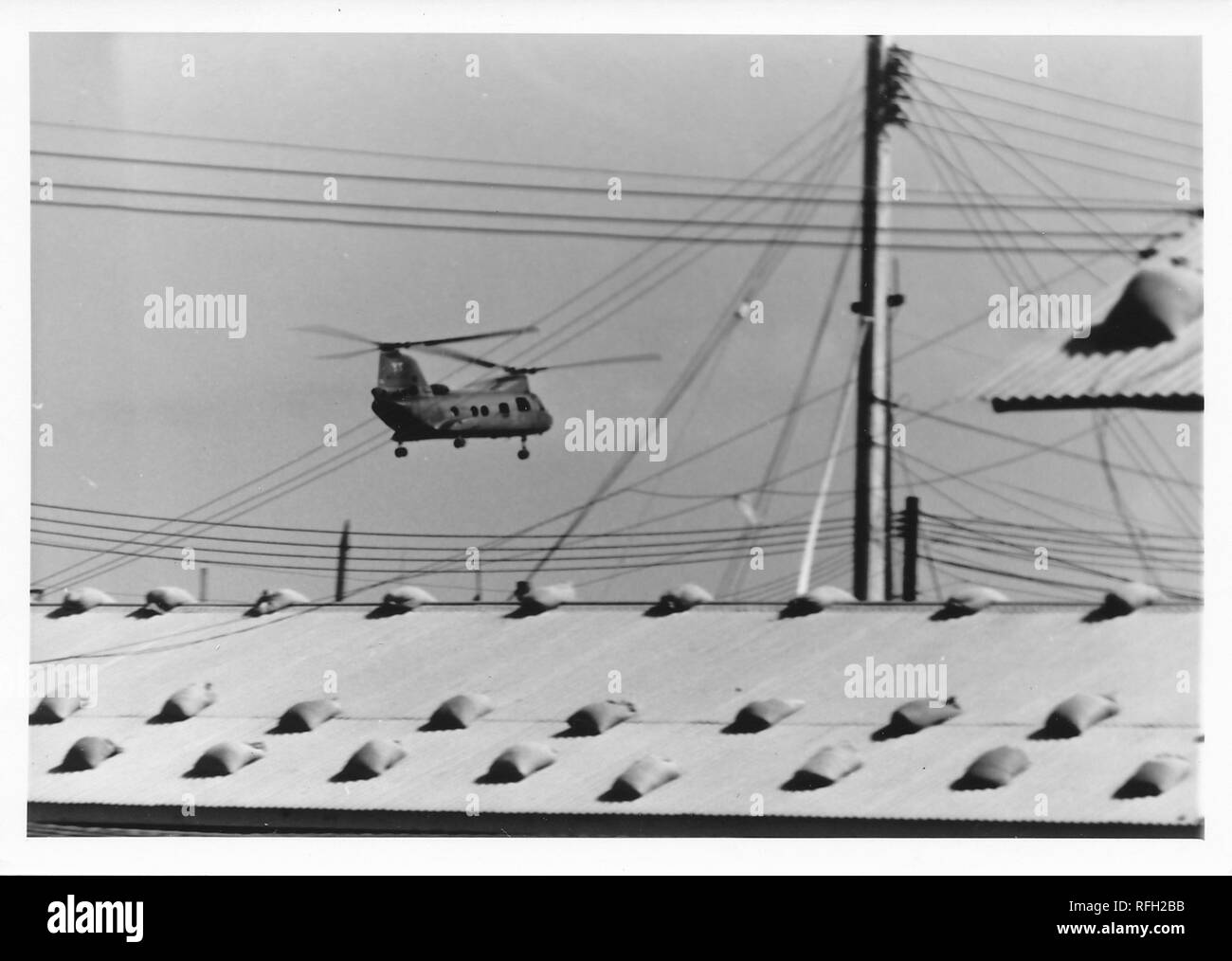 Black and white photograph, showing a tandem rotor helicopter, likely a Boeing CH-47 Chinook, in flight, with electrical wires and the roof of a barrack or administrative building visible in the foreground, photographed during the Vietnam War, 1968. () Stock Photo