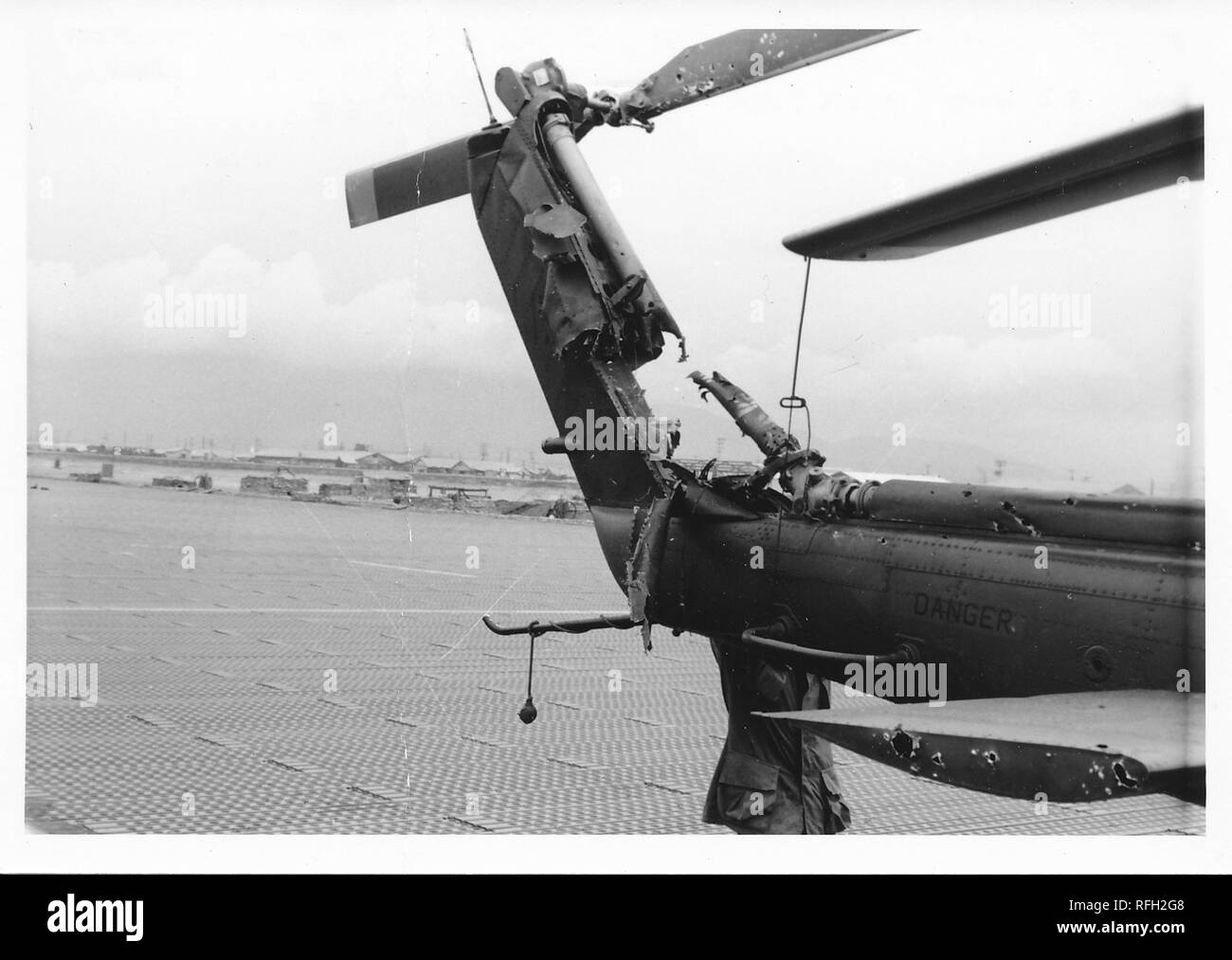 Black and white photograph, showing the heavily damaged tail of a Bell UH-1 Iroquois ('Huey') helicopter, with the trunk of a man wearing a military jacket, whose head is obscured by the tail boom, and crates and bunkers, visible in the background, photographed during the Vietnam War, 1968. () Stock Photo
