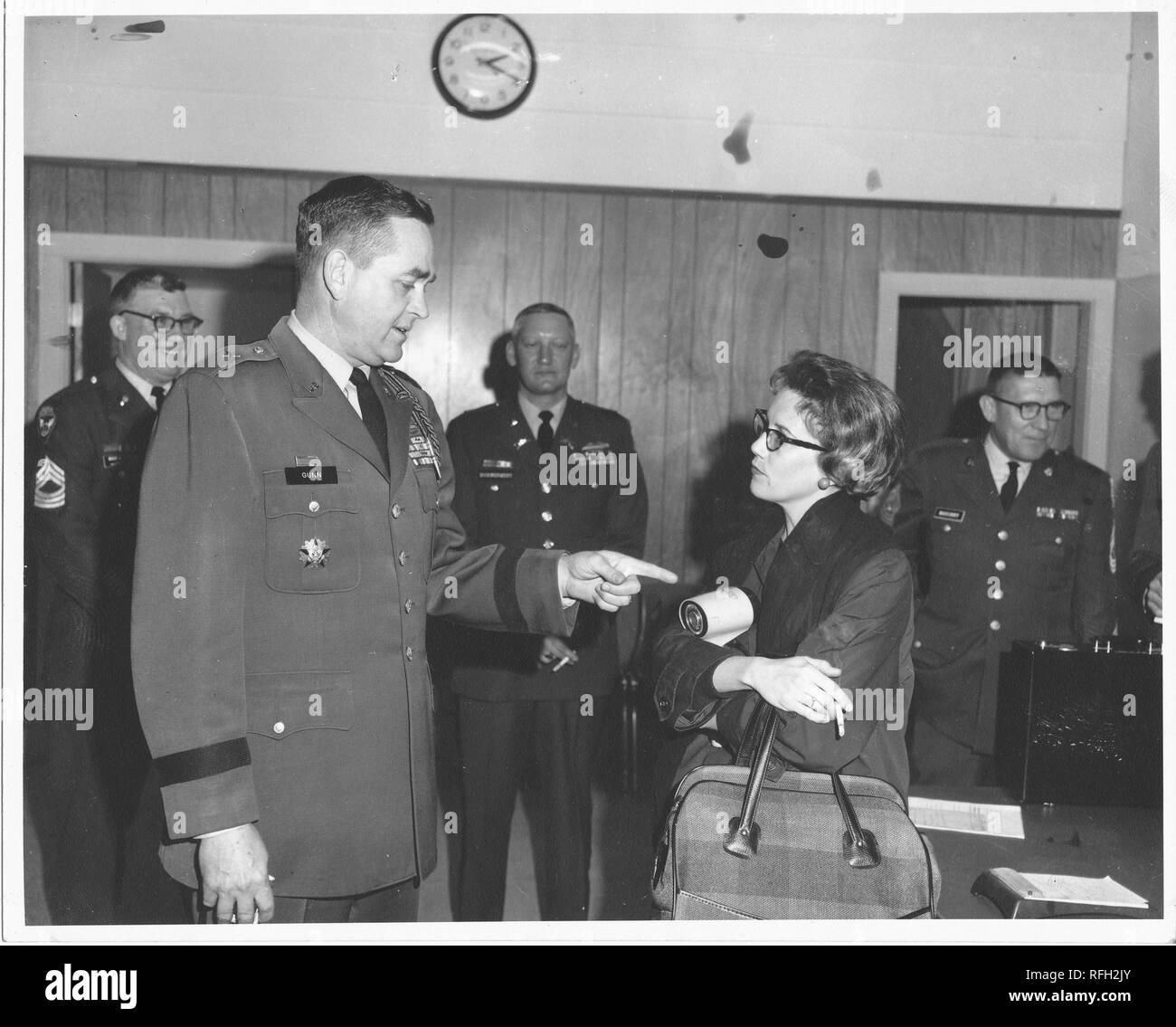 Black and white photograph of Brigadier General Frank L Gunn (left foreground) pointing to an object balanced in the crossed arms of a woman, with short dark hair, glasses, a cigarette in one hand, and a handbag draped over her arm, with several middle-aged, uniformed men, visible in the background, photographed during the Vietnam War, 1967. () Stock Photo