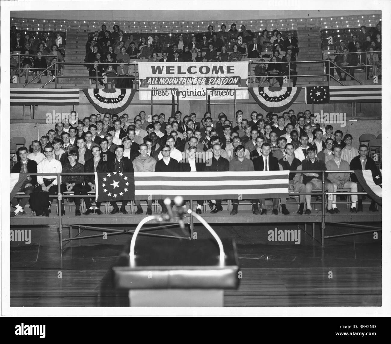 Black and white photograph of a group of young men, likely US military recruits, wearing civilian clothing, seated together on bleachers or stands in an auditorium, with older men and women seated in the upper risers, and a large sign in the background with the text 'Welcome, All Mountaineer Platoon, West Virginia's Finest, ' with a microphone visible on a podium in the foreground, photographed during the Vietnam War, 1967. () Stock Photo