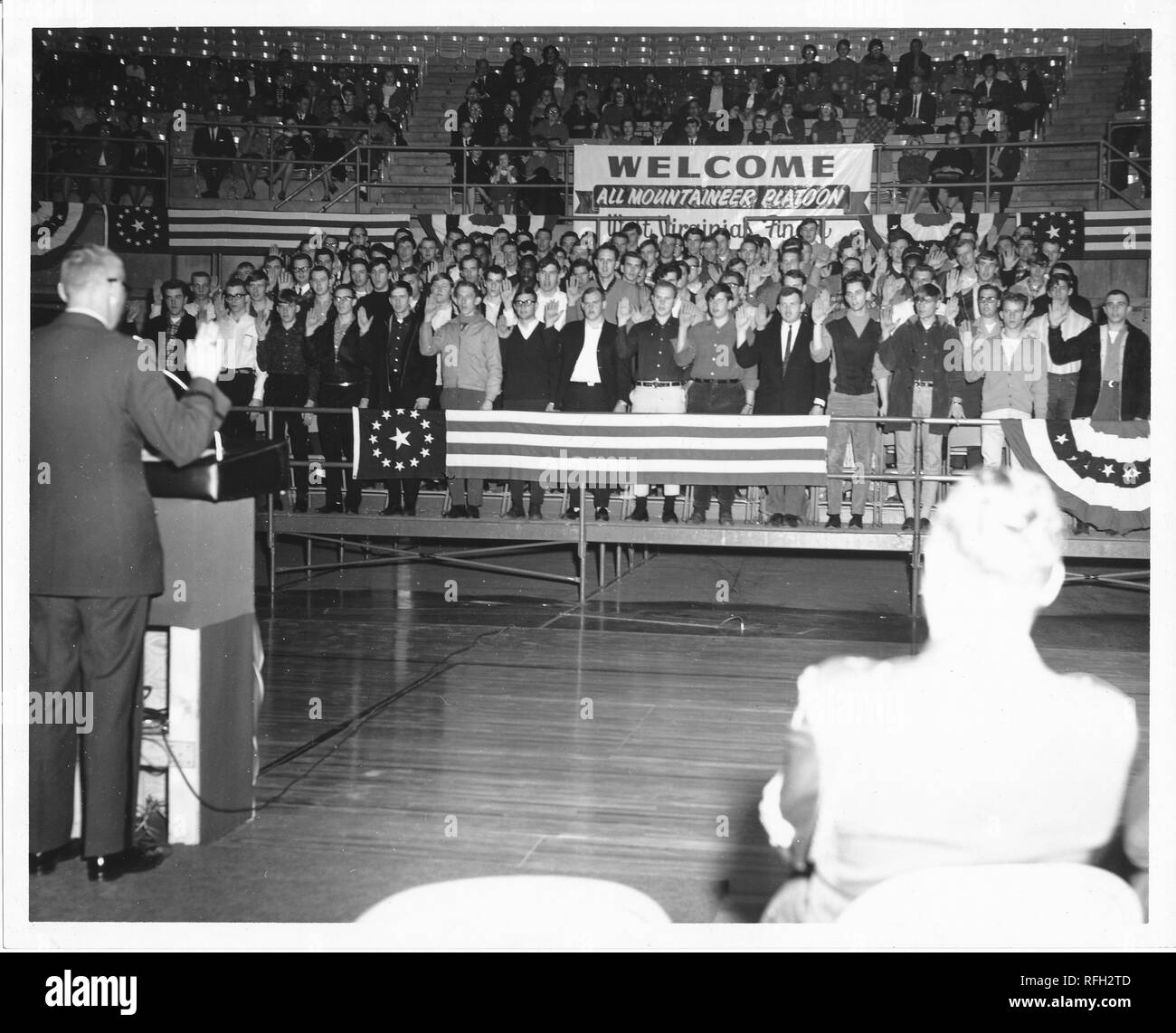 Black and white photograph of a group of young men, likely US military recruits, wearing civilian clothing, standing together on bleachers or stands in an auditorium, and raising their right hands to pledge, with older men and women seated in the upper risers, a large sign in the background with the text 'Welcome, All Mountaineer Platoon, West Virginia's Finest, ' with the back of a seated man visible in the right foreground, and a uniformed man, leading the pledge from a podium in the left foreground, photographed during the Vietnam War, 1967. () Stock Photo