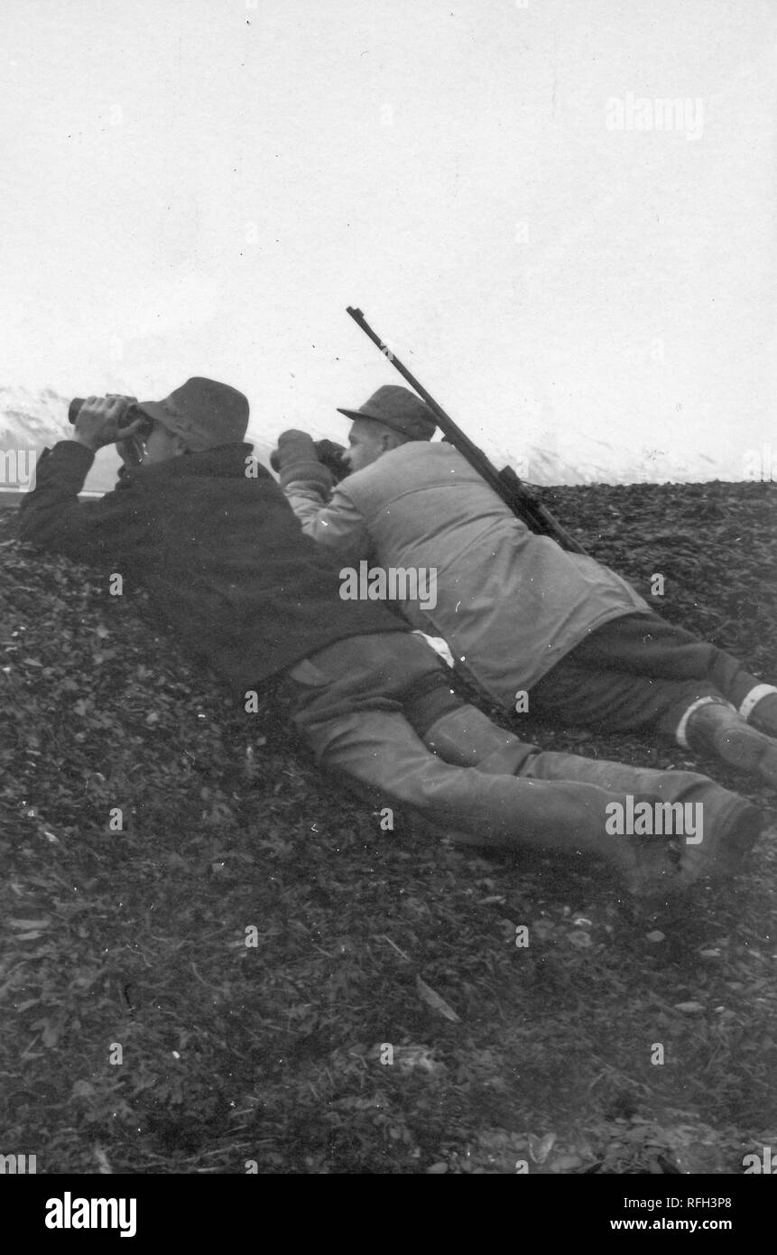 Black and white photograph of two middle-aged men wearing warm hunting jackets, boots, and hats, lying on their stomachs on the rise of a ridge and looking through binoculars, the man at right has a rifle slung over his shoulder, with snow-capped mountains visible in the background, photographed during a hunting and fishing trip located in Alaska, USA, 1955. () Stock Photo