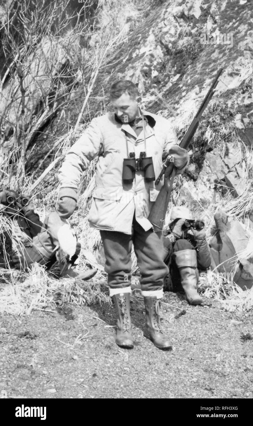 Black and white photograph of a middle-aged man standing in full-length view, wearing a hunting jacket, tall lace-up boots, and mittens, with binoculars slung around his neck and a rifle in one hand, looking down toward a dead bird held in his other hand, with two more figures behind him, each sitting on the ground and looking through binoculars, with a cliff or steep slope visible in the background, photographed during a hunting and fishing trip located in Alaska, USA, 1955. () Stock Photo