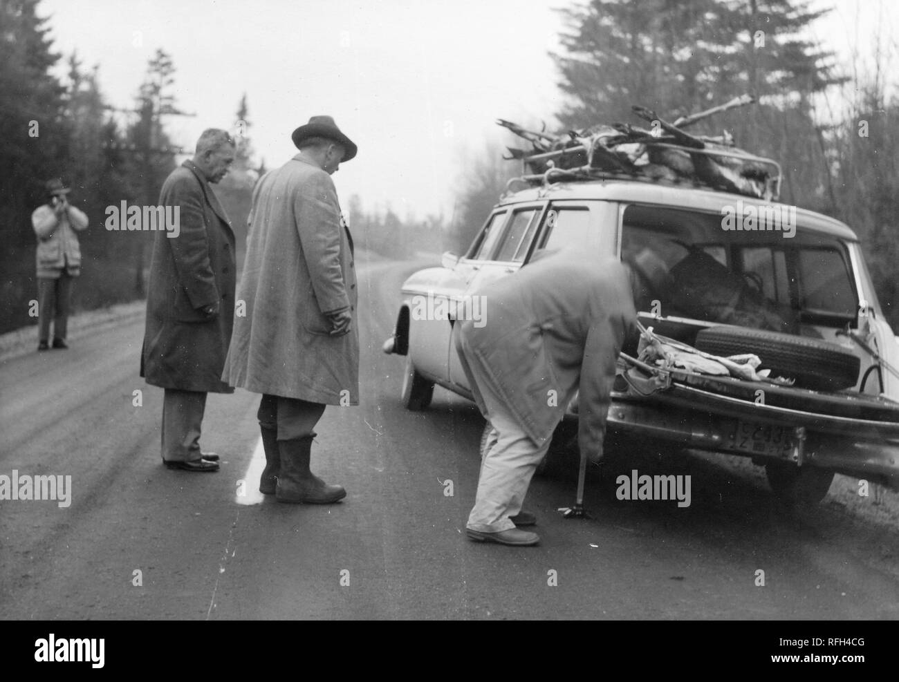 Black and white photograph of four men, each wearing a coat or jacket, standing on an empty stretch of road with a parked station wagon that has the bodies of several deer in its overhead rack; a man in the right foreground appears to change a rear tire while two men watch, the fourth man films or photographs the proceedings from the left background; with conifers and other trees visible in the background, photographed during a hunting and fishing trip located in Alaska, USA, 1955. () Stock Photo