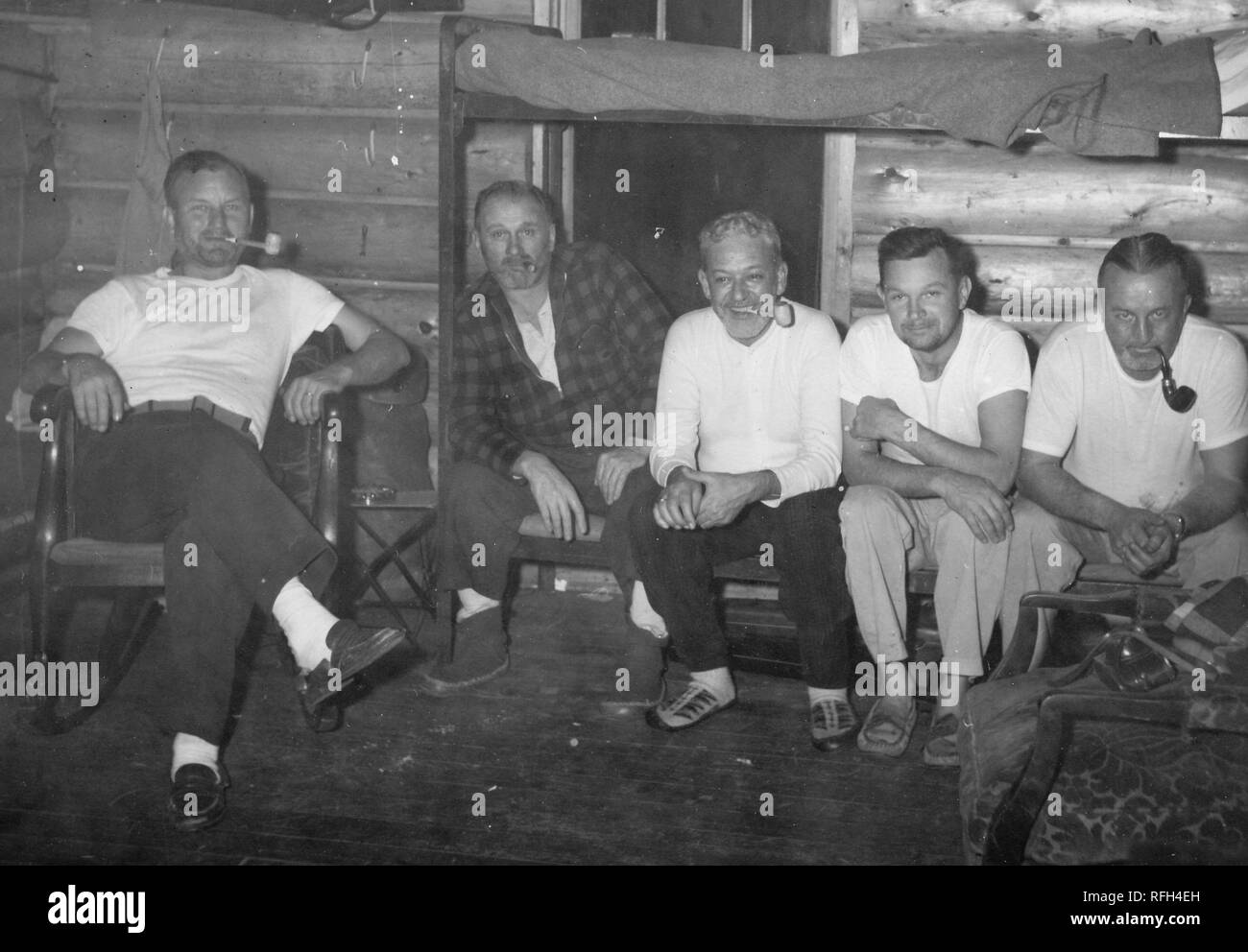 Black and white photograph of five unshaven, middle-aged men, sitting together facing the camera; one sits in a rocking chair smoking a pipe, and the other four (three smoking pipes) sit closely next to each other on the bottom bunk of a camp bed, photographed during a hunting and fishing trip located in Alaska, USA, 1955. () Stock Photo
