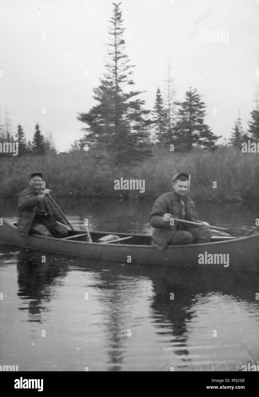 Black and white photograph of two smiling, middle-aged men, each wearing a quilted jacket and a cap, seen in three-quarter profile, seated in a canoe, with paddles in their hands; their shadow is visible in the water beneath the boat, and coniferous trees grow on the shore in the background, photographed during a hunting and fishing trip located in Alaska, USA, 1955. () Stock Photo