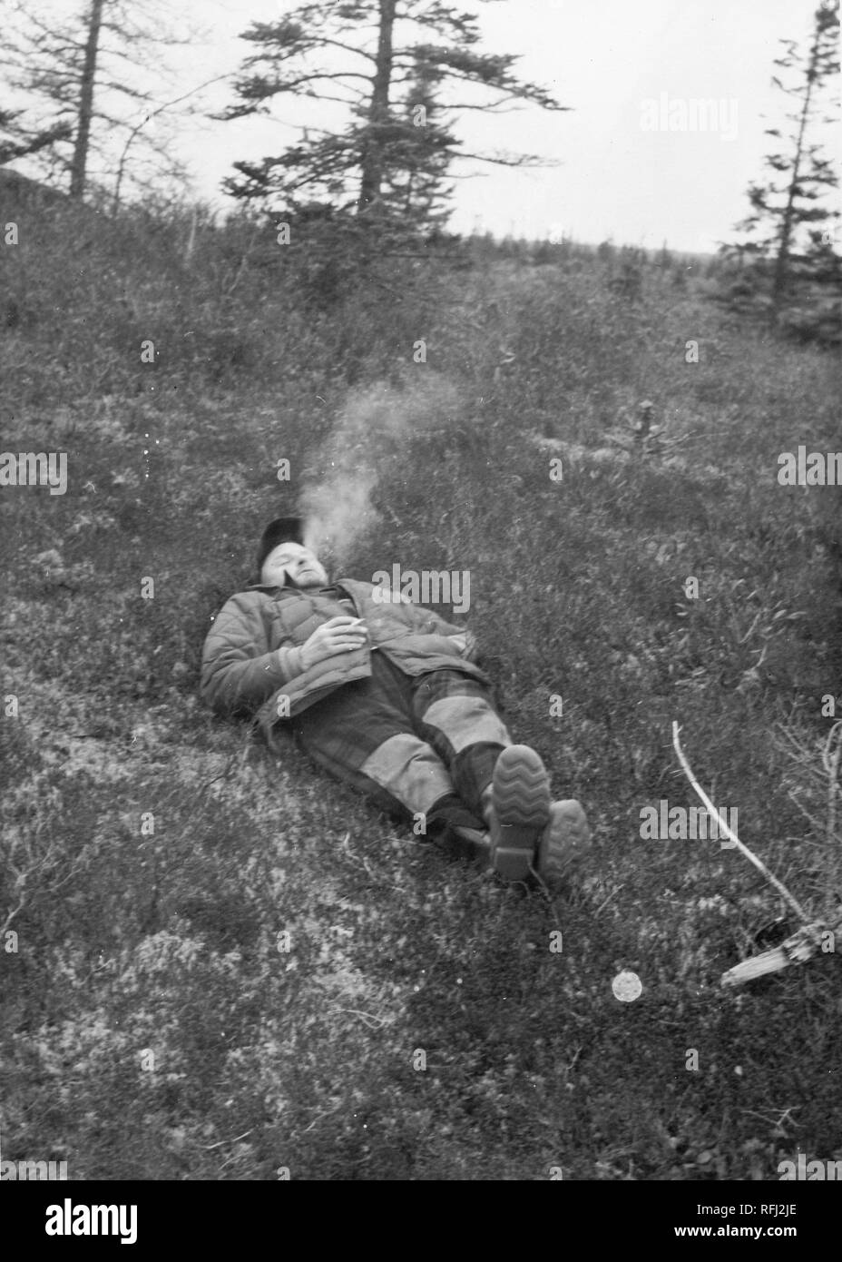 Black and white photograph of a middle-aged man, in full-length view, wearing a jacket, overalls, boots, and a cap, lying down, with his eyes closed, on a gently graded, grassy slope; one hand is in his jacket pocket, the other holds a cigarette, and recently exhaled smoke rises above his head; with several coniferous trees visible in the background, photographed during a hunting and fishing trip located in Alaska, 1955. () Stock Photo