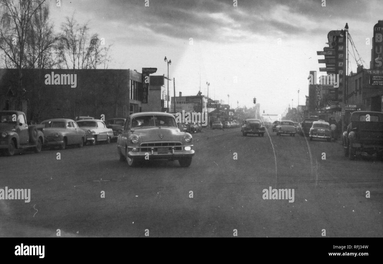 Black and white photograph of a busy main street in an Alaskan town, with vintage cars, both parked and in motion, and a long string of low buildings visible on either side of the road, with signs in the foreground advertising a 'Florist, ' the City Bus Terminal, ' an 'Emporium, ' and 'Loans, ' photographed during a hunting and fishing trip located in Alaska, 1955. () Stock Photo