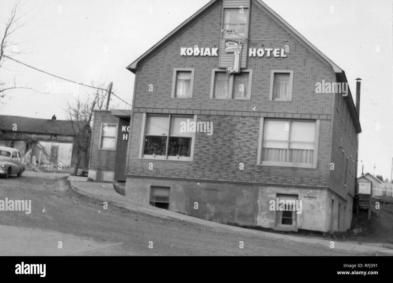 Black and white photograph shot on a slight angle, of a multi-level building with a brick facade, with the sign 'Kodiak Hotel' visible in the gable space, and several more buildings visible in the background, photographed during a hunting and fishing trip located in Alaska, 1955. () Stock Photo