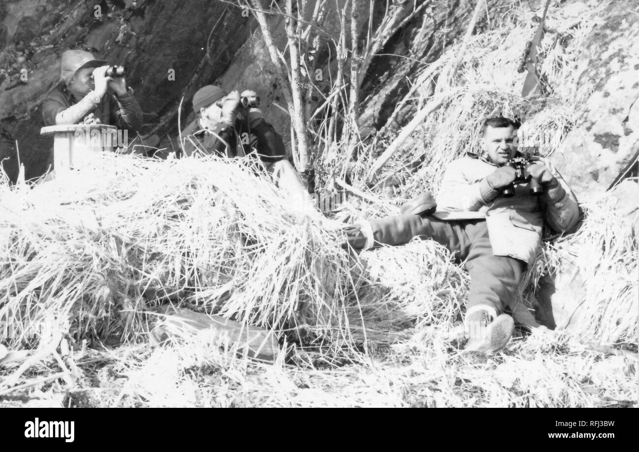 Black and white photograph of three middle-aged men in an outdoor setting with dry grasses and branches on the ground and a rocky hill or cliff in the background; two men at left use binoculars, a third man at right, wearing a warm hunting jacket, tall lace-up boots, and mittens, reclines on the ground holding his binoculars away from his face, photographed during a hunting and fishing trip located in Alaska, 1955. () Stock Photo